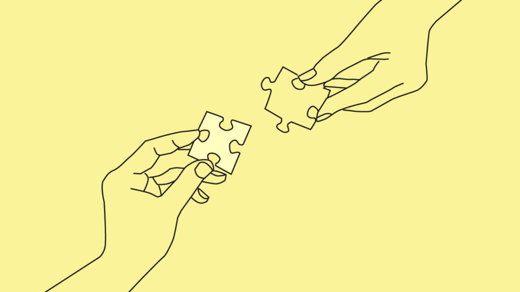 Grafika przedstawia dwie ręce trzymające elementy puzzli, które są w trakcie łączenia. Ręce są narysowane czarnymi liniami na żółtym tle. Elementy puzzli są białe i mają charakterystyczny kształt z wypustkami i wcięciami.