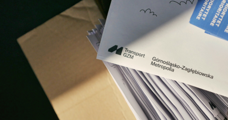 Zdjęcie przedstawia stos białych kopert z nadrukowanym logo Transport GZM i napisem "Górnośląsko-Zagłębiowska Metropolia". Na kopertach widoczne są również niebieskie naklejki z napisem "PRIORYTET" w kilku językach. W lewym dolnym rogu kadru widać fragment kartonowego pudełka.